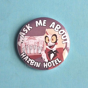 Puede incluir: Una chapa redonda con un personaje de dibujos animados de pelo rubio, traje rojo y pajarita. El texto "ASK ME ABOUT HAZBIN HOTEL" está impreso en blanco alrededor del borde. El fondo muestra una escena urbana estilizada con figuras.