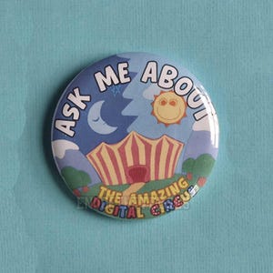 Puede incluir: Una chapa redonda con una colorida ilustración de una carpa de circo a rayas bajo una luna y un sol. El texto ASK ME ABOUT THE AMAZING DIGITAL CIRCUS aparece en letras negritas. El fondo muestra montañas azules y árboles verdes.