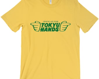 Puede incluir: Una camiseta amarilla con un gráfico verde que dice "CREATIVE LIFE STORE TOKYU HANDS" con dos manos verdes apuntando hacia la izquierda y la derecha.
