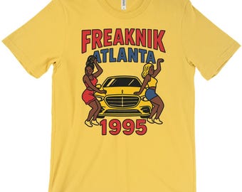 Puede incluir: Camiseta amarilla mostaza con el texto "FREAKNIK ATLANTA 1995" en rojo y azul. El gráfico presenta dos figuras bailando y un coche amarillo. La camiseta está hecha de un material suave y tiene un cuello redondo clásico.