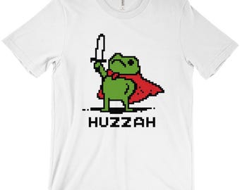 Puede incluir: Camiseta blanca con una rana verde pixelada que lleva una capa roja y sostiene una espada. La palabra "Huzzah" está impresa en letras negras debajo.