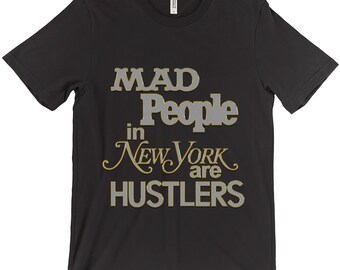 Puede incluir: Camiseta negra con el texto "MAD People in NEW YORK are HUSTLERS" en una fuente estilizada. El texto está en gris con un contorno dorado.