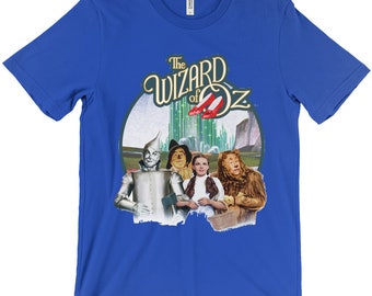 Puede incluir: Una camiseta azul real con un gráfico de estilo vintage de los personajes de El Mago de Oz. El gráfico presenta a Dorothy, el Espantapájaros, el Hombre de Hojalata y el León Cobarde de pie frente a la Ciudad Esmeralda. El texto "The Wizard of Oz" está escrito en una fuente estilizada sobre los personajes.
