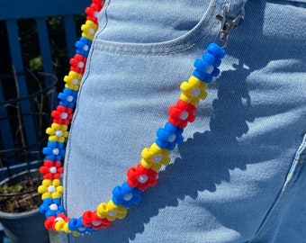 May include: A beaded chain made of blue, red, yellow, and white plastic flowers. The chain is attached to a silver clip and is hanging from a pair of blue jeans.