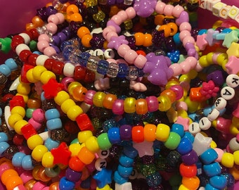 Könnte beinhalten: Ein rosa Eimer gefüllt mit bunten Perlenarmbändern. Die Armbänder sind aus verschiedenen Farben von Perlen gefertigt, darunter Regenbogen, Rosa, Lila, Gelb, Orange, Rot, Blau, Grün, Schwarz und Weiß. Einige der Armbänder haben sternförmige Perlen, und einige haben Buchstabenperlen, die Wörter wie "Kid" und "Kailey" buchstabieren.