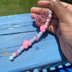 May include: A pink and white beaded keychain with pink heart charms and a clear plastic charm. The keychain is held in a hand.