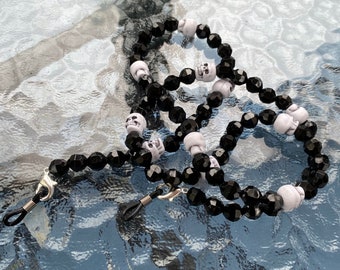 May include: Eyeglass chain featuring black faceted beads and white skull beads. The chain has silver-colored clasps for attaching to glasses. A unique accessory with a gothic theme.