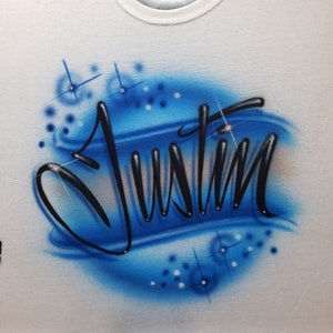 May include: A white t-shirt with a blue and white airbrushed design featuring the name "Justin" in black lettering.