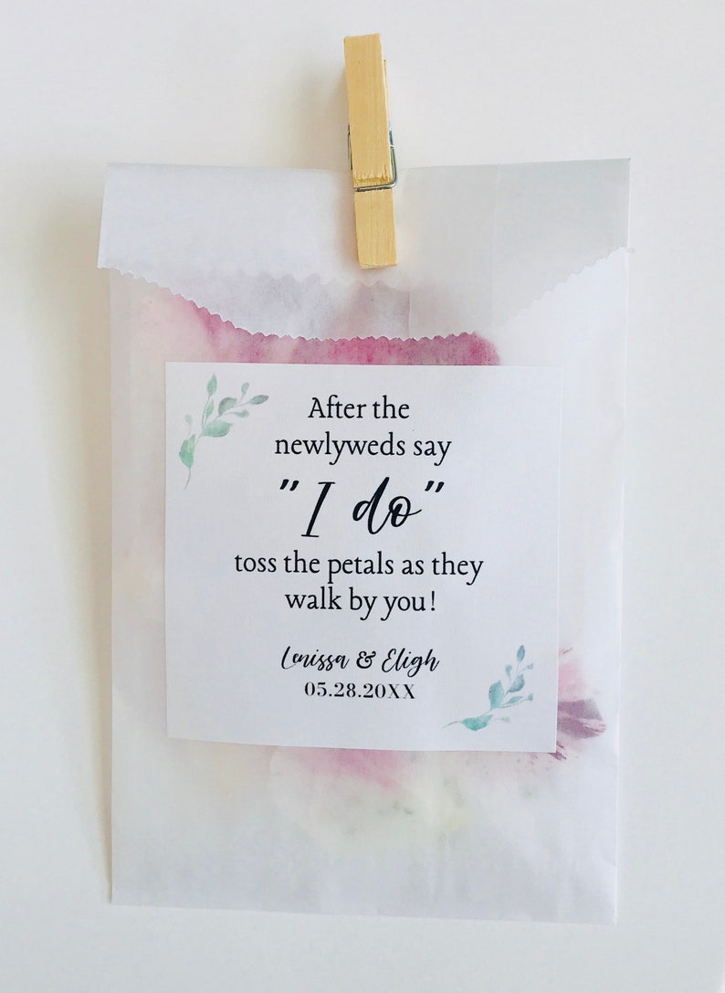 Puede incluir: Una bolsa de papel blanca con un dise&ntilde;o de acuarela rosa y verde. La bolsa tiene una etiqueta que dice "Despu&eacute;s de que los reci&eacute;n casados digan "S&iacute;, quiero" tira los p&eacute;talos mientras pasan por ti!" y "Lenissa & Eligh 05.28.20XX".
