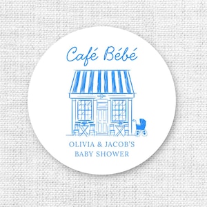 Puede incluir: Una pegatina redonda y blanca con una ilustraci&oacute;n azul de un caf&eacute;, un cochecito de beb&eacute; y el texto "Caf&eacute; B&eacute;b&eacute;" y "OLIVIA & JACOB'S BABY SHOWER". El caf&eacute; tiene un toldo a rayas y mesas y sillas peque&ntilde;as.