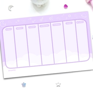 May include: A lavender weekly planner with a whimsical design featuring stars, a crescent moon, and a planet. The planner has seven columns for each day of the week, with the days of the week labeled at the top. The planner has a section at the bottom labeled "dreams:".