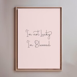 May include: A framed print featuring the text "I'm not lucky, I'm Blessed" in black cursive on a pale pink backdrop. The frame is a warm brown, and the print is mounted on a neutral grey wall.