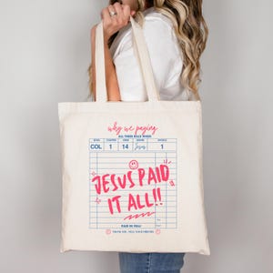 May include: A cream-colored tote bag with a pink and blue graphic design. The text on the bag reads "Why we paying all these bills when Jesus paid it all!!" The bag has two handles and is being held by a person.