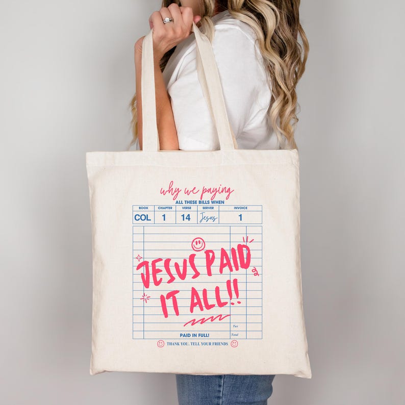 May include: A cream-colored tote bag with a pink and blue graphic design. The text on the bag reads "Why we paying all these bills when Jesus paid it all!!" The bag has two handles and is being held by a person.