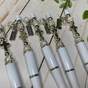 May include: A collection of white pens with silver accents and cross charms. Each pen features a decorative top with rhinestones and a dangling tag that reads "Faith". The pens are arranged on a light wood surface.