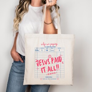 May include: Cream-colored tote bag with the text "JESUS PAID IT ALL!!" in pink, designed to resemble a bill. The bag also features the phrases "why we paying" and "PAID IN FULL!" in a handwritten style. The bag has a simple, minimalist design.