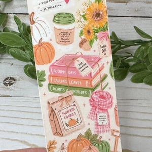 May include: A rectangular bookmark featuring autumn-themed illustrations including pumpkins, sunflowers, a coffee cup, a stack of books labeled Autumn Days, Falling Leaves, and Hearts & Hayrides, a bag of pumpkin cookies, a scarf, and a wooden wagon.
