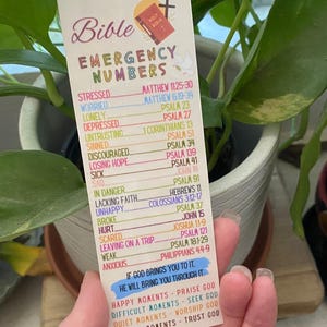 May include: A bookmark with the words "Bible Emergency Numbers" and a list of Bible verses for various emotional states. The bookmark has a cross and a small illustration of a book. The bottom of the bookmark has a blue banner with a quote.