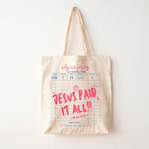 May include: Cream-colored tote bag with a graphic design resembling a bill. The text "JESUS PAID IT ALL!!" is in bright pink, with additional text above and below. The bag has long handles for carrying.