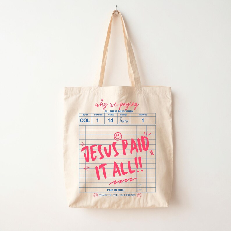 May include: Cream-colored tote bag with a graphic design resembling a bill. The text "JESUS PAID IT ALL!!" is in bright pink, with additional text above and below. The bag has long handles for carrying.