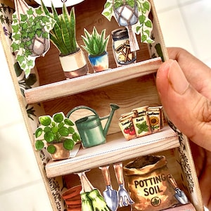May include: A miniature three-tiered wooden display shelf featuring paper cutouts of gardening supplies. The top shelf displays hanging plants, potted succulents, and a jar. The middle shelf shows a watering can and seed packets. The bottom shelf holds gardening gloves, tools, and a bag labeled POTTING SOIL.