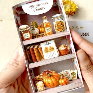 Può includere: Un mobiletto in carta in miniatura intitolato The Autumn Cabinet, con tre ripiani decorati con illustrazioni di conserva di mele, sidro, fette di arancia essiccata, libri, un calendario di ottobre, una tazza e zucche.