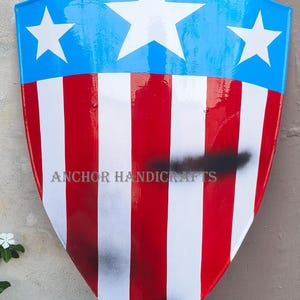 Puede incluir: Artículo decorativo en forma de escudo con una sección superior azul con tres estrellas blancas. La sección inferior tiene rayas verticales rojas y blancas. El texto "ANCHOR HANDICRAFTS" es visible. Un diseño patriótico.