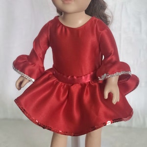 May include: A doll with brown hair and green eyes wearing a red satin dress with a flared skirt and bell sleeves. The dress is trimmed with silver sequins. A pink bow adorns the doll's hair.