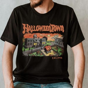 A black t-shirt with a Halloween-themed graphic design depicting a spooky town scene with a jack-o-lantern, buildings, and a cemetery.