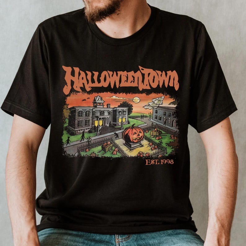 A black t-shirt with a Halloween-themed graphic design depicting a spooky town scene with a jack-o-lantern, buildings, and a cemetery.