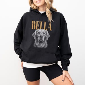A woman wearing a black hoodie with a graphic of a dog's face and the name "Bella" printed on it.