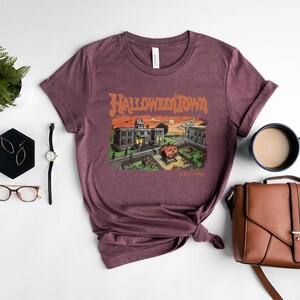 a purple t-shirt with a Halloween-themed graphic, a pair of glasses, a watch, a coffee cup, a handbag, and a potted plant arranged on a white background.