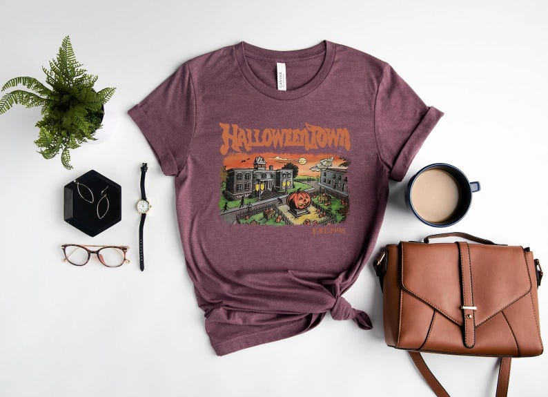 a purple t-shirt with a Halloween-themed graphic, a pair of glasses, a watch, a coffee cup, a handbag, and a potted plant arranged on a white background.