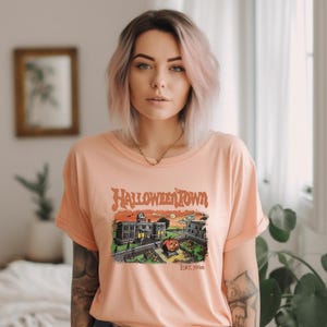 A young woman with pink hair wearing a t-shirt with a Halloween-themed graphic stands in a room with a window and plants in the background.