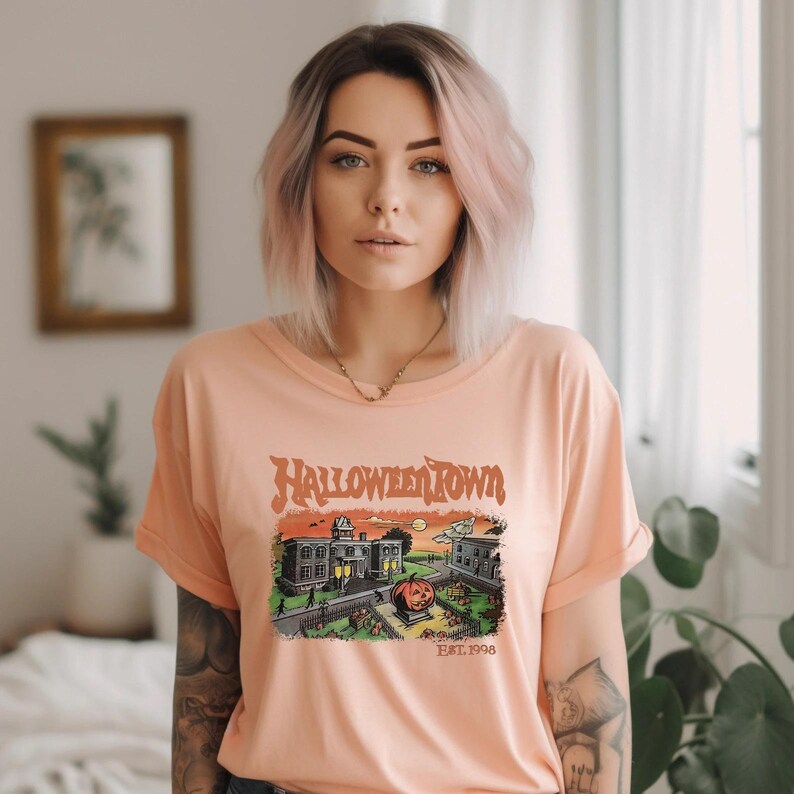A young woman with pink hair wearing a t-shirt with a Halloween-themed graphic stands in a room with a window and plants in the background.