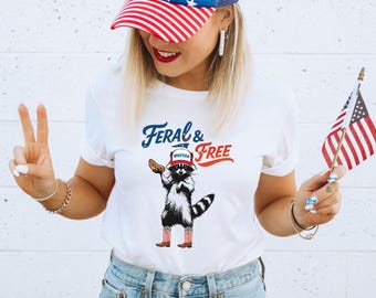 May include: White t-shirt with the text "Feral & Free" and a raccoon graphic wearing a hat that says "America" and holding a hot dog. The person is wearing a red, white, and blue baseball cap and holding a small American flag.