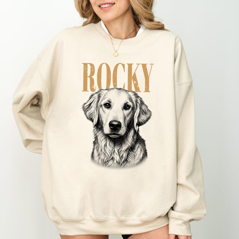 A woman wearing a beige sweatshirt with a graphic of Rocky, a dog, printed on it.
