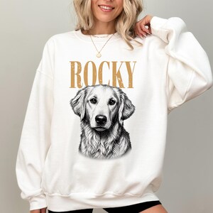 A woman wearing a white sweatshirt with a graphic of a dog's face and the word "ROCKY" printed on it.