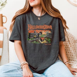 A woman wearing a t-shirt with a graphic of a Halloween-themed town, sitting on a couch.