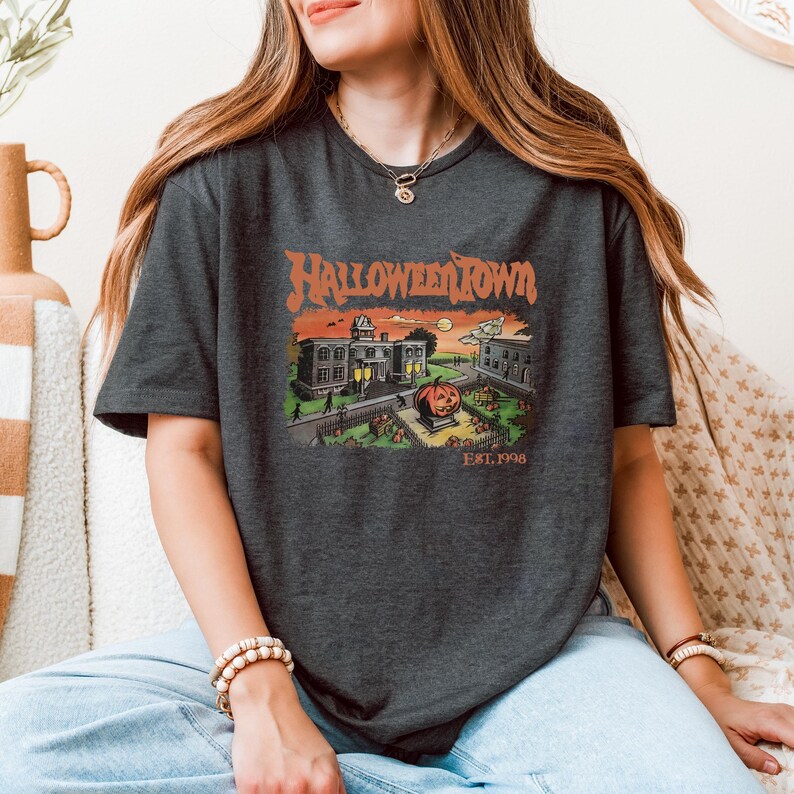A woman wearing a t-shirt with a graphic of a Halloween-themed town, sitting on a couch.