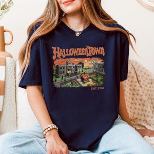 A woman wearing a blue t-shirt with a Halloween-themed graphic design, sitting on a couch.