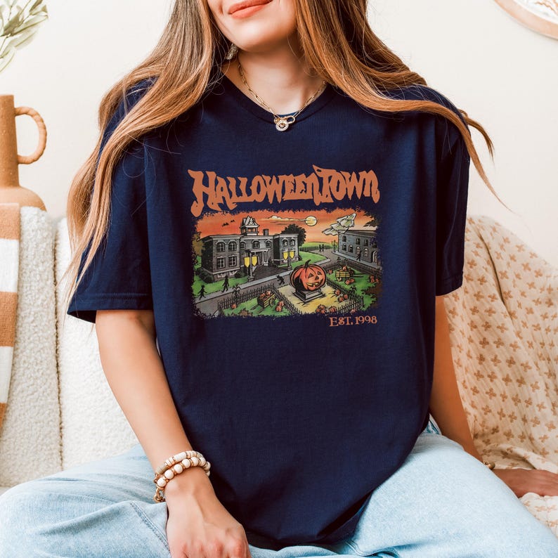 A woman wearing a blue t-shirt with a Halloween-themed graphic design, sitting on a couch.