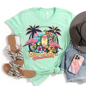 May include: A light green t-shirt with a graphic of two pink flamingos, palm trees, and a tropical drink. The text "Five O'Clock Somewhere" is printed in a retro font.
