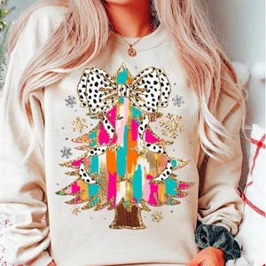 May include: A cream-colored sweatshirt with a colorful Christmas tree design. The tree is decorated with a large black and white polka dot bow, gold glitter, and colorful stripes.