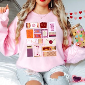 Peut inclure: Un sweat-shirt rose clair orné d'un collage de boîtes d'allumettes, de cartes et d'enveloppes illustrées. Les motifs incluent les mots "Lucky You" et "Love", ainsi que d'autres graphiques. Le sweat-shirt est de style décontracté.