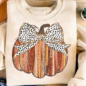 May include: A beige sweatshirt featuring a graphic of a pumpkin with a black and white polka dot bow. The pumpkin is orange and brown with a striped pattern.