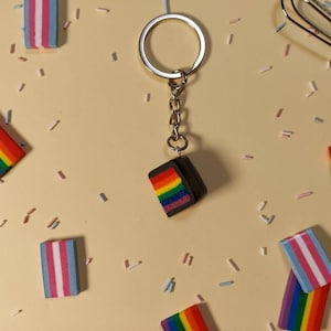 May include: A rainbow pride flag cake keychain with a silver key ring. The keychain is surrounded by rainbow and transgender pride flag cake charms on a light beige surface.