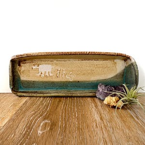 May include: A long, rectangular ceramic serving dish with a light brown glaze and a turquoise blue glaze at the bottom. The dish has a white design of a moose and people walking, with a cross symbol.
