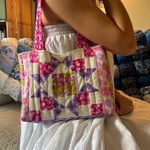 May include: A quilted tote bag featuring a patchwork star pattern in shades of pink, purple, white, and olive green. The bag has two matching pink floral fabric handles and a rectangular body constructed from various floral and geometric fabric squares.