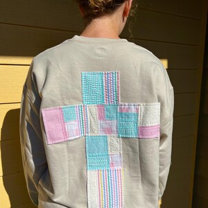 May include: A beige sweatshirt with a patchwork cross design on the back. The cross is made of various pastel-colored fabrics, including pink gingham, blue seersucker, and striped patterns. The sweatshirt is a casual, comfortable garment.